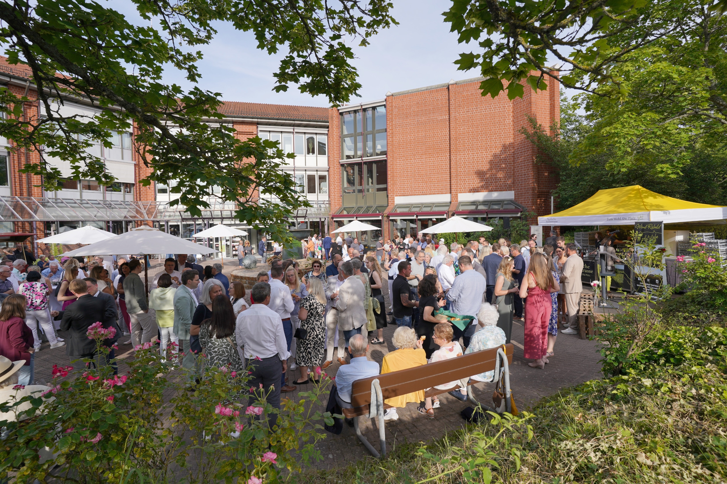 Eine Outdoor-Veranstaltung auf dem Platz vor dem Kreishaus der Kreisverwaltung Südliche Weinstraße, der Kreisempfang am 8. Juni 2024. Menschen stehen unter weißen Sonnenschirmen und um ein gelbes Zelt vor dem roten Backsteingebäude der Kreisverwaltung. Das Wetter ist sonnig und die Veranstaltung ist gut besucht, mit einer vielfältigen Gruppe von Menschen, die sich unterhalten und das Event genießen.