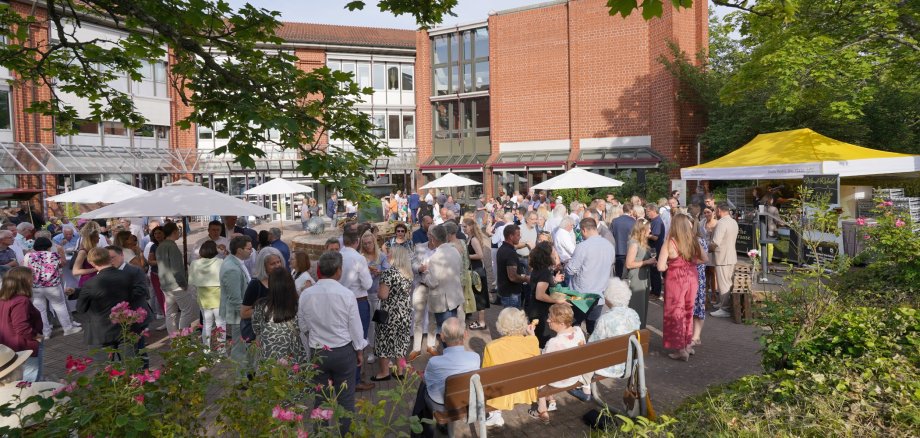 Eine Outdoor-Veranstaltung auf dem Platz vor dem Kreishaus der Kreisverwaltung Südliche Weinstraße, der Kreisempfang am 8. Juni 2024. Menschen stehen unter weißen Sonnenschirmen und um ein gelbes Zelt vor dem roten Backsteingebäude der Kreisverwaltung. Das Wetter ist sonnig und die Veranstaltung ist gut besucht, mit einer vielfältigen Gruppe von Menschen, die sich unterhalten und das Event genießen.