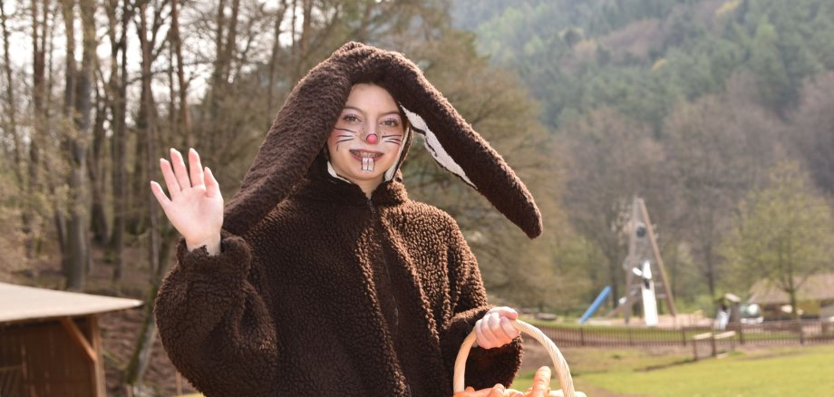 Frau im Osterhasenkostüm trägt einen Korb mit Karotten und winkt in die Kamera.