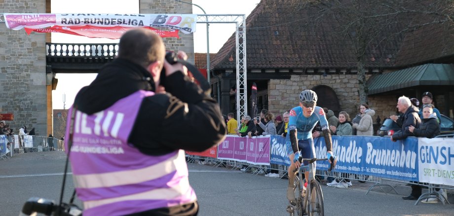 Ein Fotograf und Publikum vor dem Deutschen Weintor, durch das Radfahrer ins Ziel kommen,