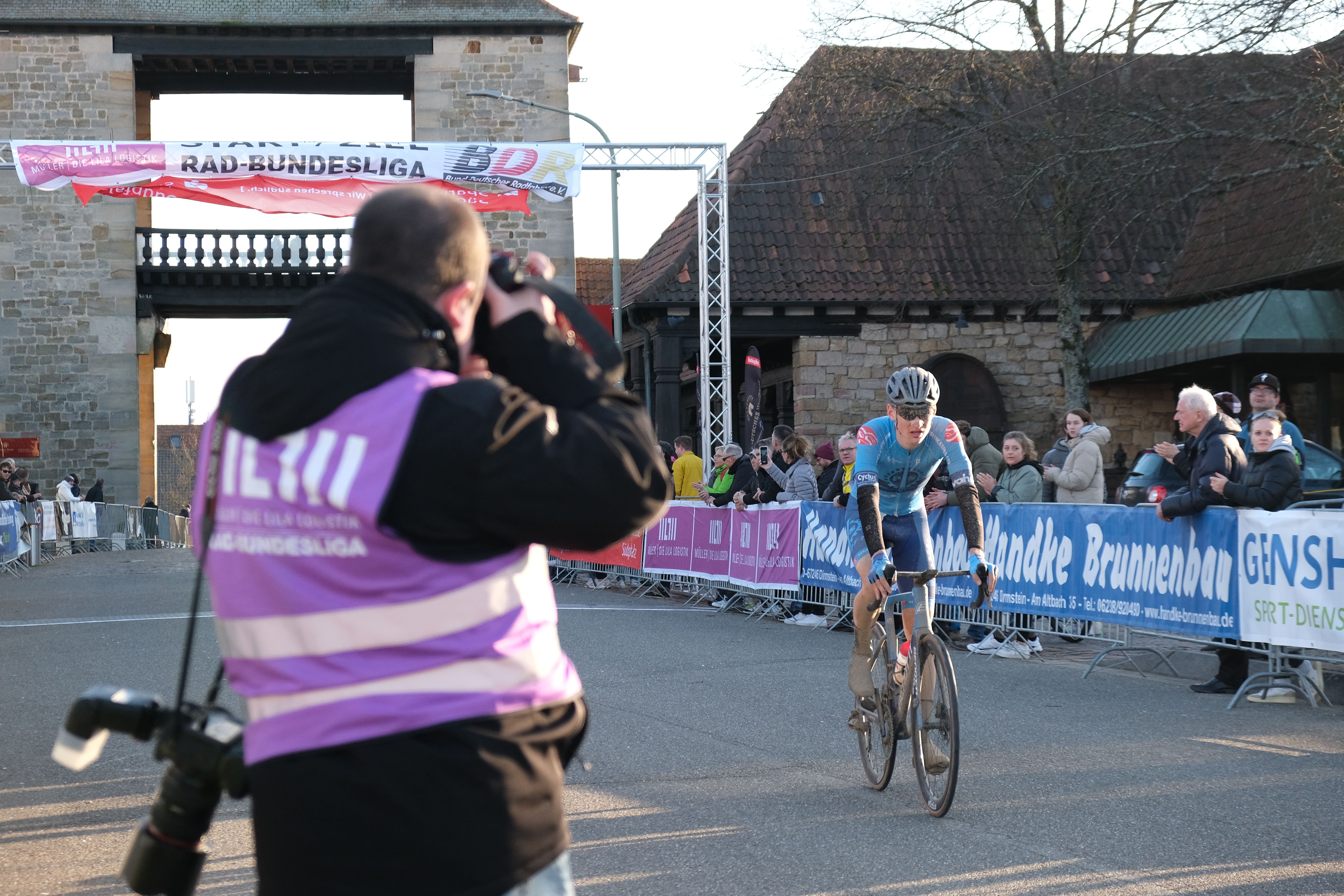 Ein Fotograf und Publikum vor dem Deutschen Weintor, durch das Radfahrer ins Ziel kommen,