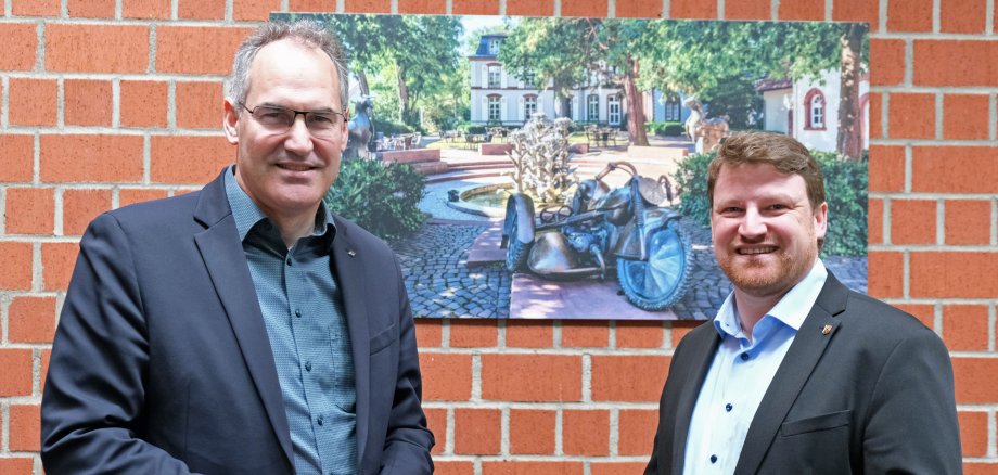 Ortsbürgermeister Sven Koch und Landrat Dietmar Seefeldt stehen vor einem Bild im Kreishaus, das den Tabakbrunnen in Herxheim zeigt.