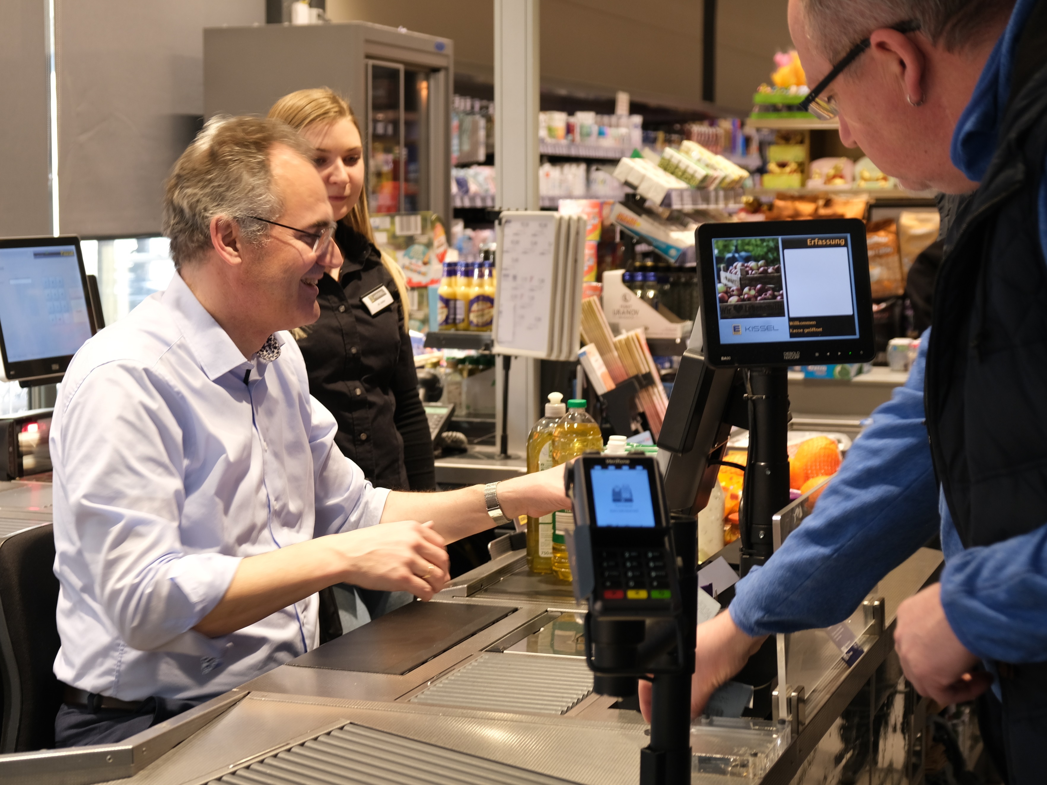 Landrat Dietmar Seefeldt sitzt an einer Supermarktkasse und bedient einen Kunden. 