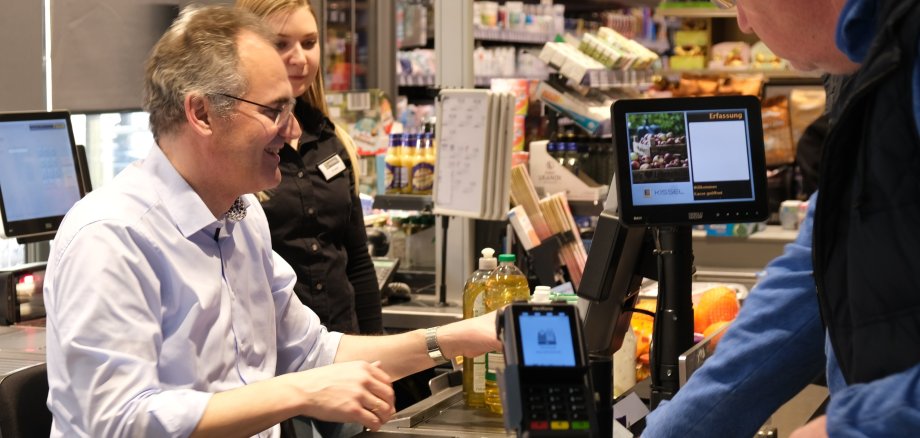 Landrat Dietmar Seefeldt sitzt an einer Supermarktkasse und bedient einen Kunden. 