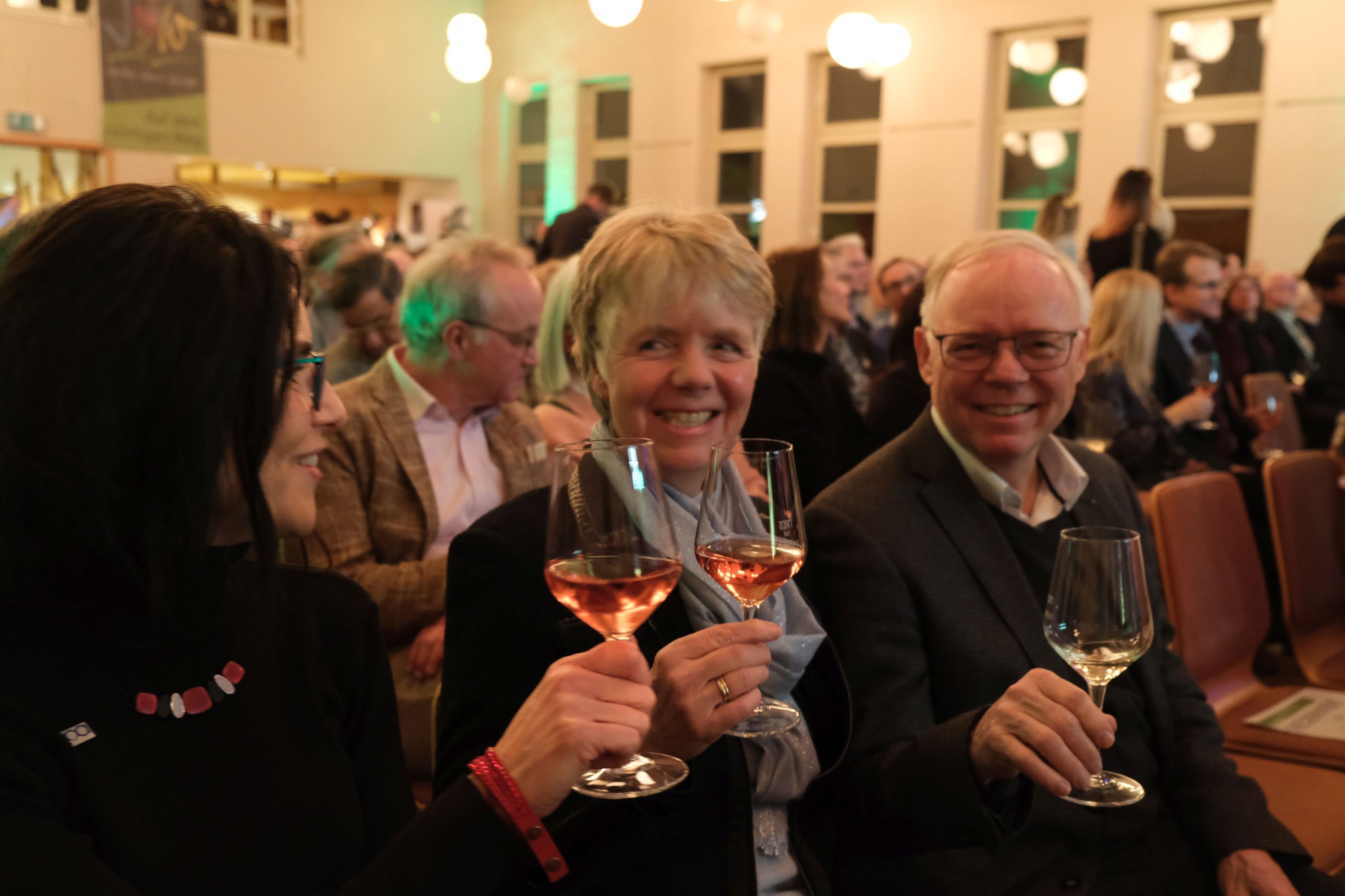 Professor Dr. Reinhard Töpfer stößt mit einem Glas Sekt beim diesjährigen Neujahrsempfang des Landkreises Südliche Weinstraße in Billigheim-Ingenheim mit zwei Frauen an. Im Hintergrund sitzen viele Menschen im bestuhlten Saal des Bürgerhauses. Foto: KV SÜW