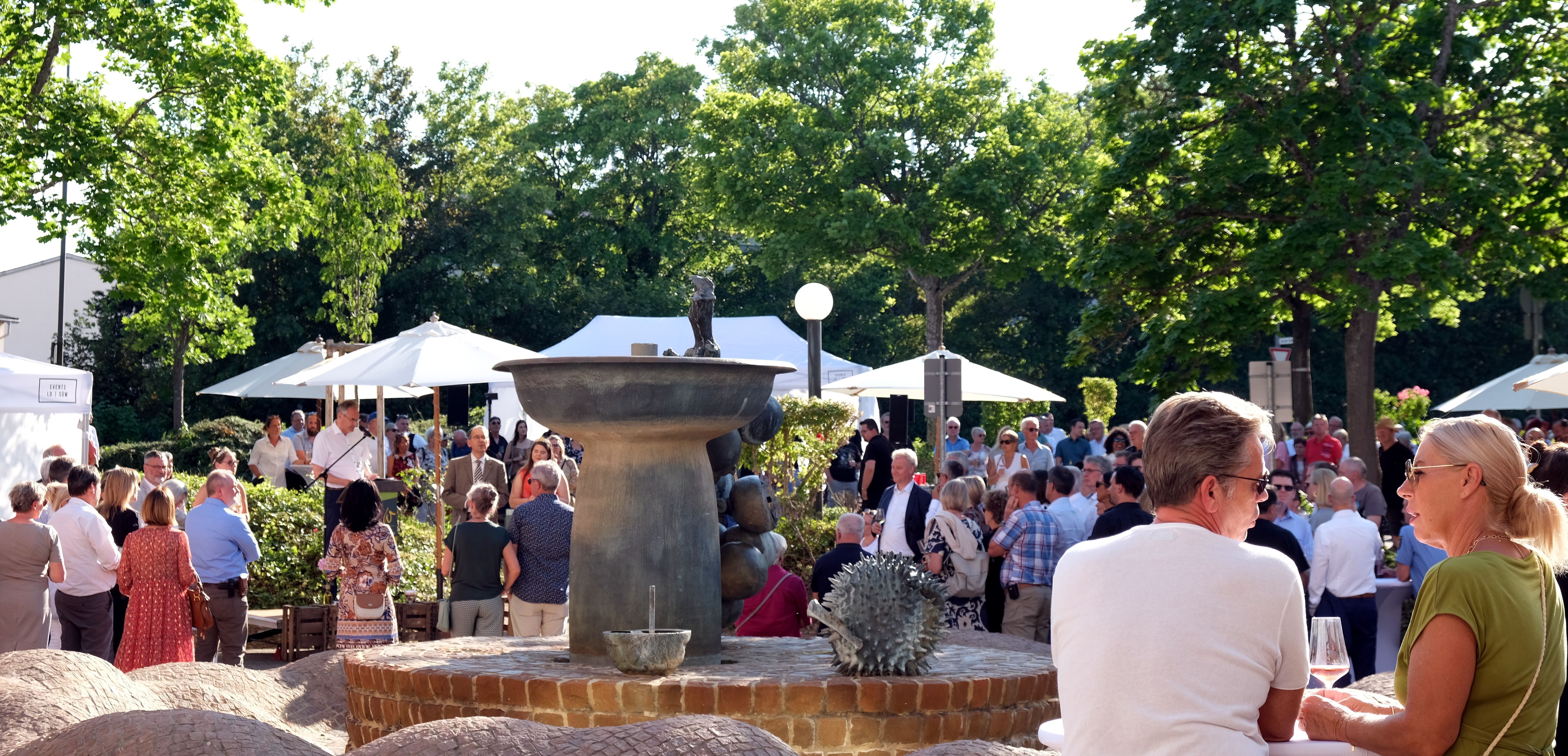 Rings um den sogenannten Früchtebrunnen vor dem Kreishaus in Landau stehen viele Menschen, die Landrat Dietmar Seefeldt beim Vortragen seiner Ansprache zuhören. Im Vordergrund eine Frau und ein Mann, die sich unterhalten.