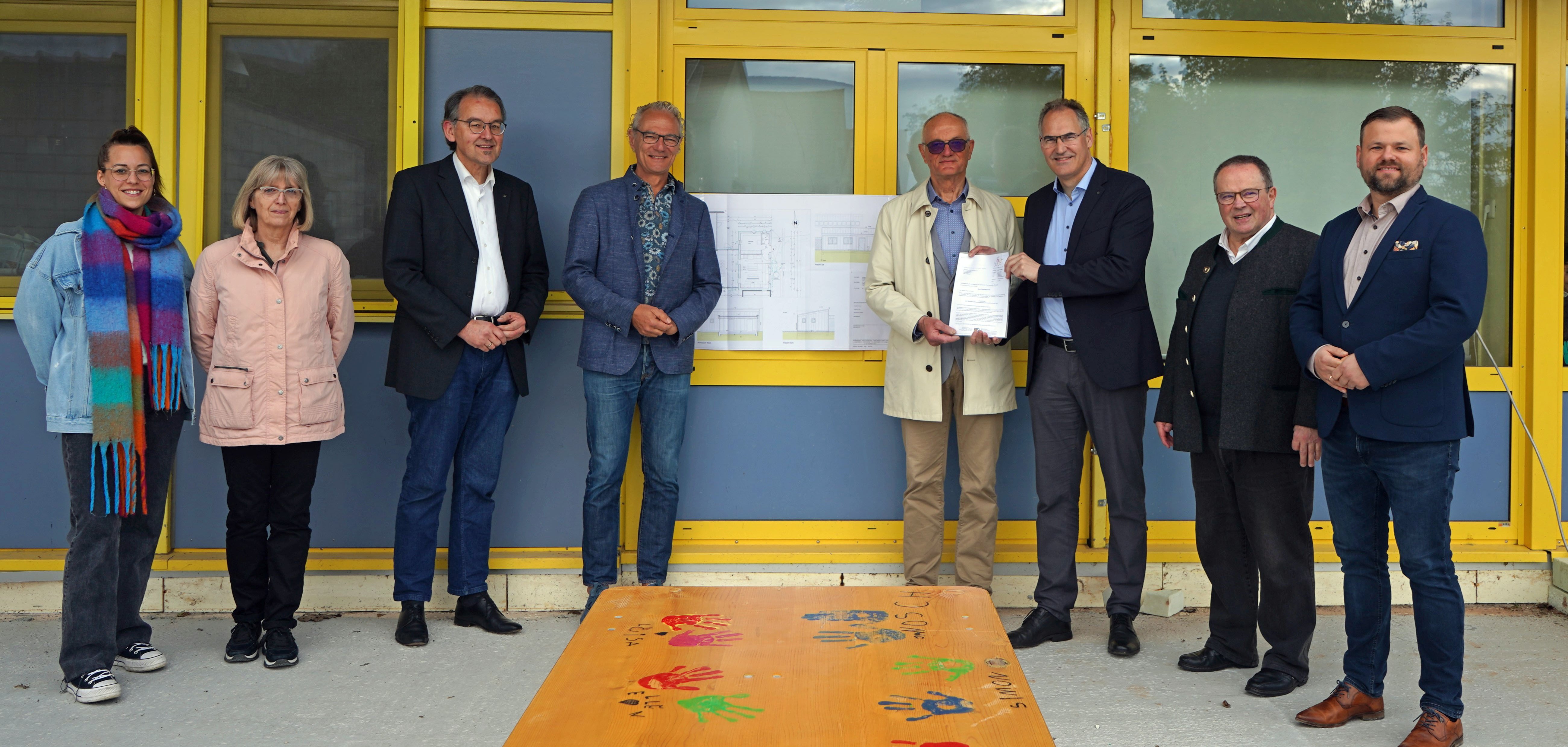 Eine Gruppe von acht Personen steht vor einem Gebäude mit gelben Fenstern und blauen Wänden. Vor ihnen ist ein Tisch mit bunten Handabdrücken von Kindern. Die Gruppe besteht aus Lisa Bullinger, Vertreterin des Elternausschusses, Michaela Esser, Leiterin der Kita St. Josef, Erster Kreisbeigeordneter Georg Kern, Offenbachs Ortsbürgermeister Axel Wassyl, Lothar Bibus vom St. Elisabethenverein, Landrat Dietmar Seefeldt, Pfarrer Arno Vogt von der Pfarrei Heiliger Laurentius Herxheim und Offenbachs Beigeordnetem Simon Wingerter.