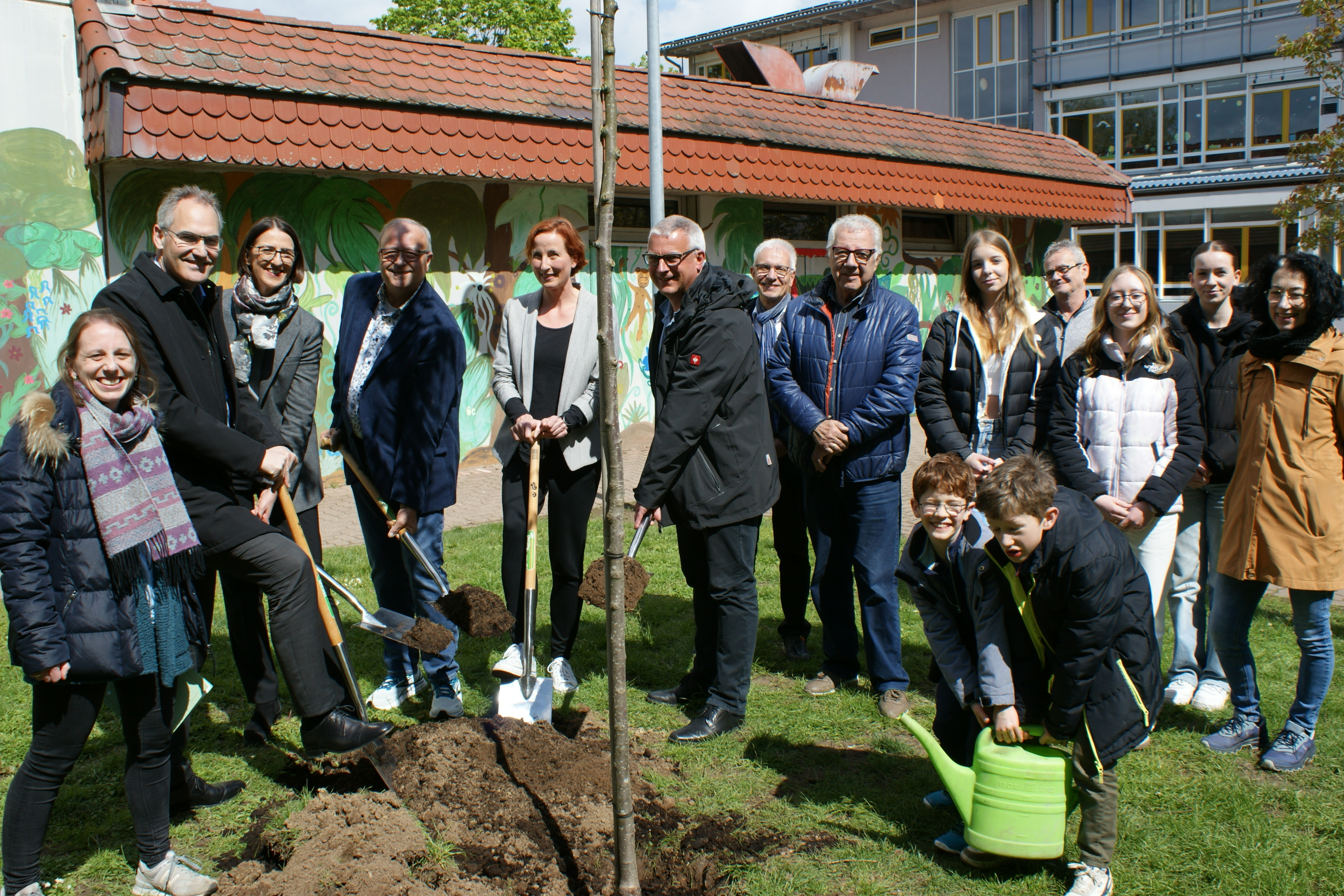 Vor dem PAMINA-Schulzentrum in Herxheim stehen Landrat Dietmar Seefeldt und zahlreiche weitere Personen - teils mit Schaufel in der Hand - , um anlässlich des Tags des Baumes 2024 einen Mehlbeerbaum zu pflanzen. Im Vordergrund tragen zwei junge Schüler gemeinsam eine Gießkanne, um den frisch gepflanzten Baum zu wässern.