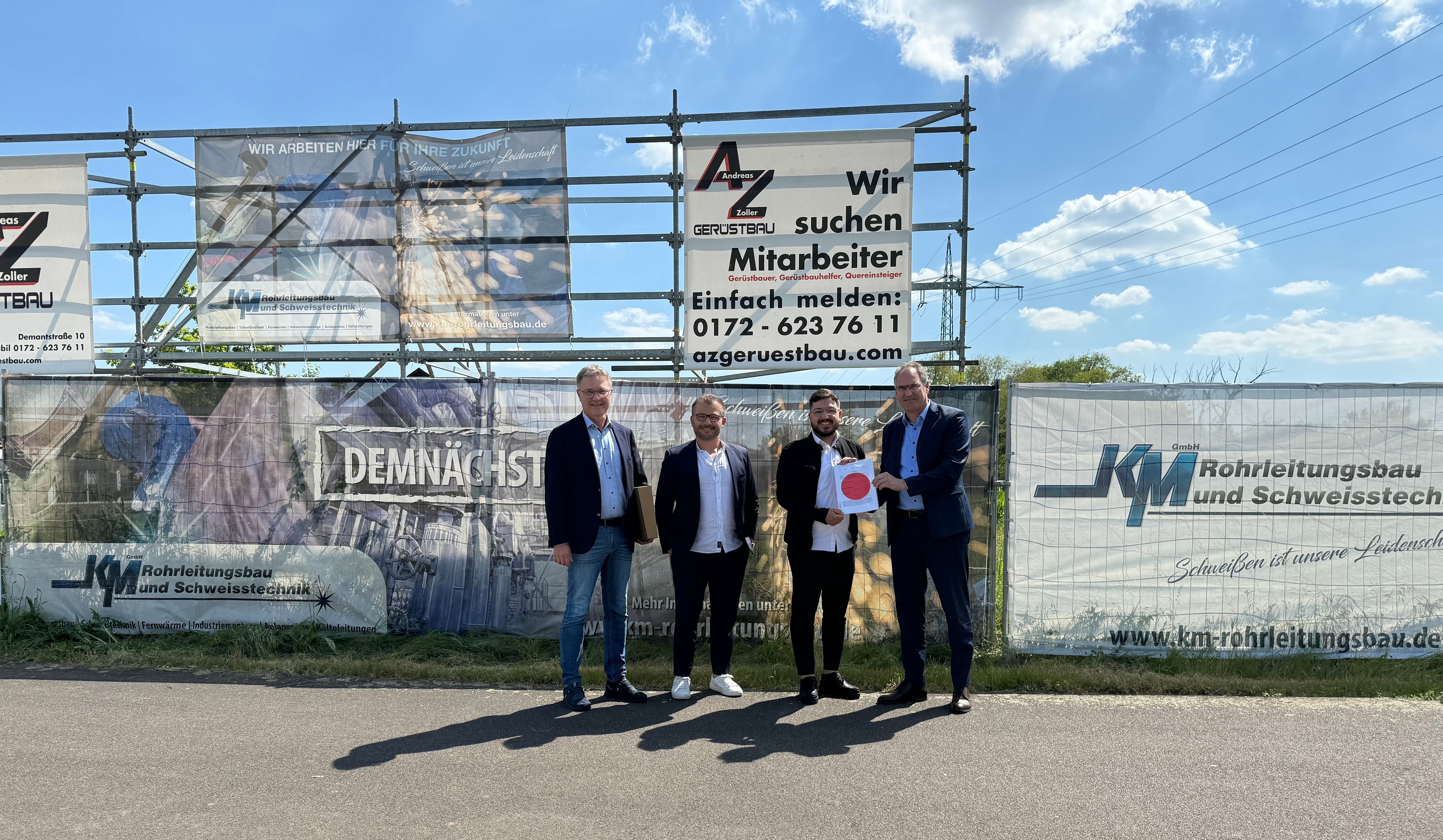 Landrat Dietmar Seefeldt (rechts) übergibt die Baugenehmigung mit einem großen roten Punkt darauf zur offiziellen Freigabe des Bauprojekts im Beisein von Günter Jung, Leiter der Abteilung Bauen und Umwelt bei der Kreisverwaltung (links), an den Geschäftsführer und Gesellschafter Adem Karamese (Zweiter von rechts) und Projektleiter Tarkan Üncü der KM Rohrleitungsbau und Schweisstechnik GmbH. Die vier stehen vor der Baustelle bzw. einem Bauzaun.