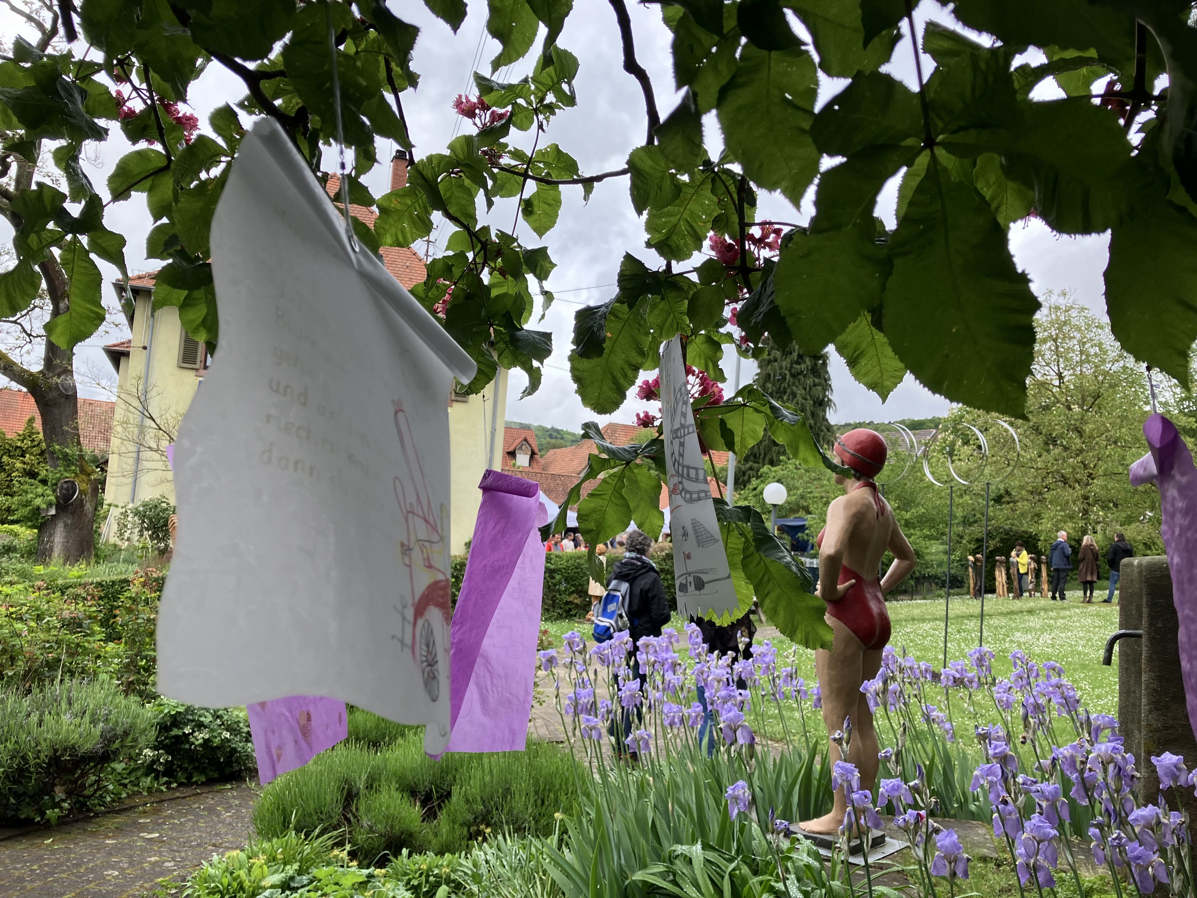 Bunte Zeichnungen und Texte hängen an einer Schnur, die zwischen Bäumen im Garten gespannt ist. Im Hintergrund steht eine lebensgroße Skulptur einer Frau im Badeanzug und mit Badekappe, die "stehende Schwimmerin". Weiter im Hintergrund ist eine hohe Stahlskulptur namens „Clouds“ zu sehen. Menschen spazieren im Garten.