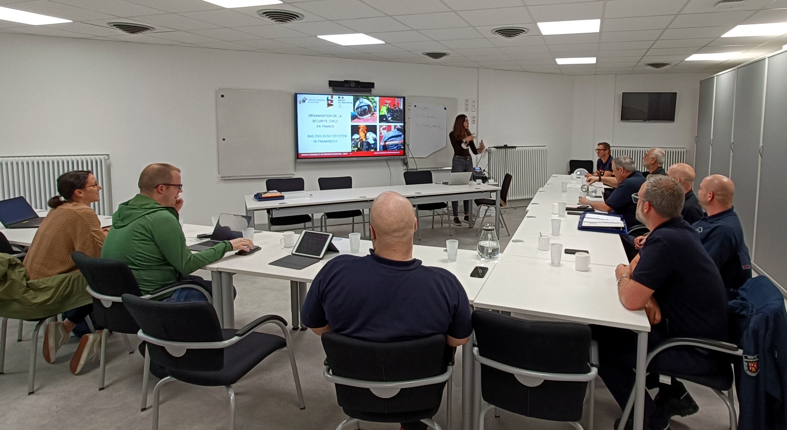 Eine Gruppe von Vertreterinnen und Vertretern des Katastrophenschutzes der Südlichen Weinstraße und des französischen Departements Bas-Rhin sitzt an einem Konferenztisch in einem modernen Sitzungsraum. Eine Frau hält eine Präsentation vor einem Bildschirm, auf dem Informationen zur Organisation des Katastrophenschutzes in Frankreich angezeigt werden.