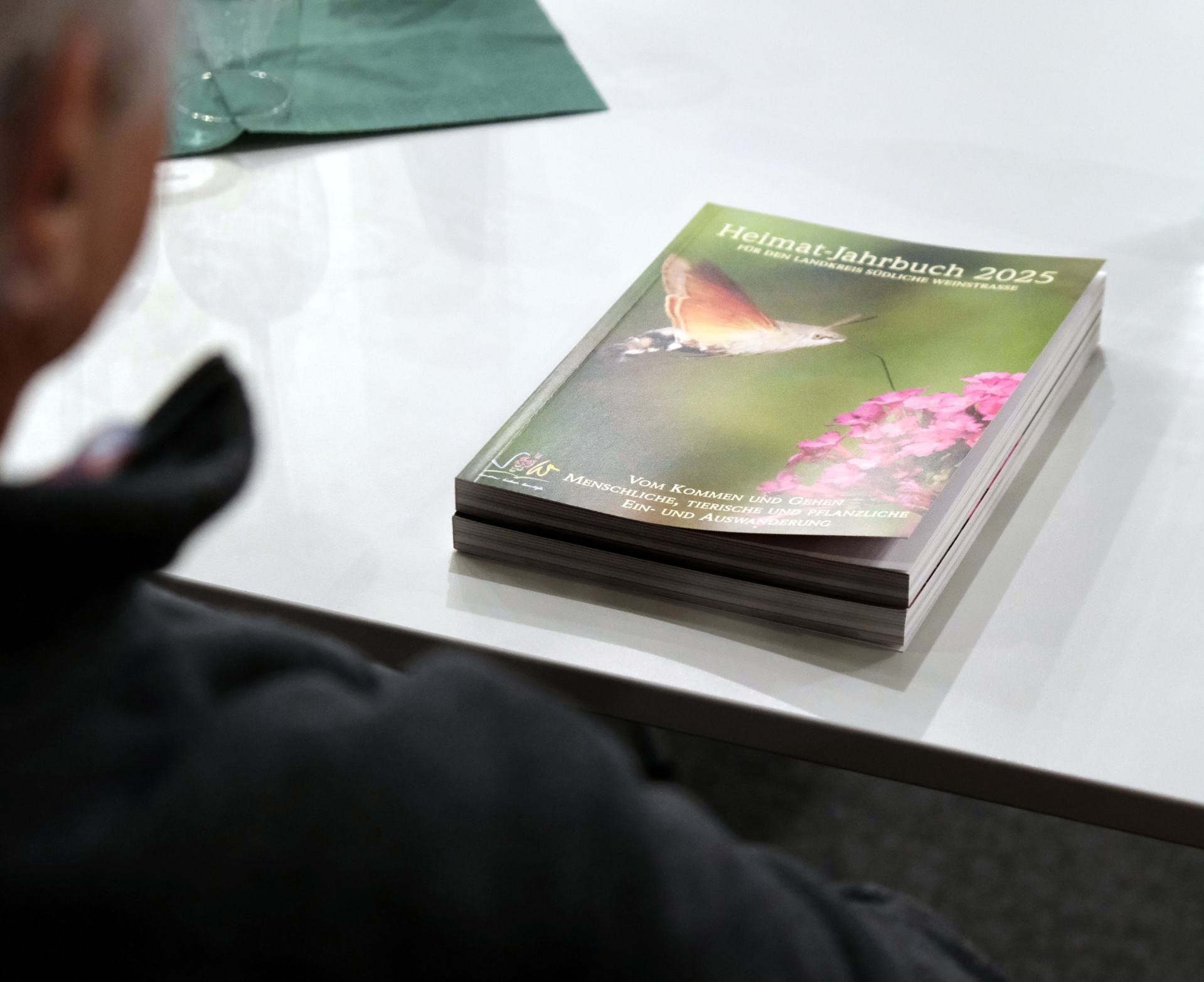 Blick über die Schulter eines Lesers auf das neue Heimatjahrbuch, das auf einem Tisch liegt. Das Titelfoto des Buches zeigt ein Taubenschwänzchen beim Anflug auf einen Sommerflieder.