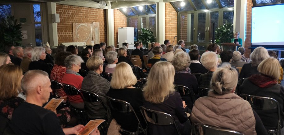 Eine größere Gruppe von Menschen sitzt im Casino der Kreisverwaltung und hört einem Vortrag zu. Im Vordergrund ist das Publikum von hinten zu sehen, während Gleichstellungsbeauftragte Isabelle Stähle am Rednerpult vorne rechts spricht.