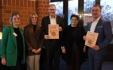 Gruppenfoto beim Festakt für STOPP: Die Gleichstellungsbeauftragten Isabelle Stähle (SÜW), Laura Hess (LD), Lisa-Marie Trog sowie Landrat Dietmar Seefeldt (SÜW) und Christoph Buttweiler, Erster Kreisbeigeordneter GER, halten ein Plakat mit der Einladung zur Veranstaltung (von links nach rechts).