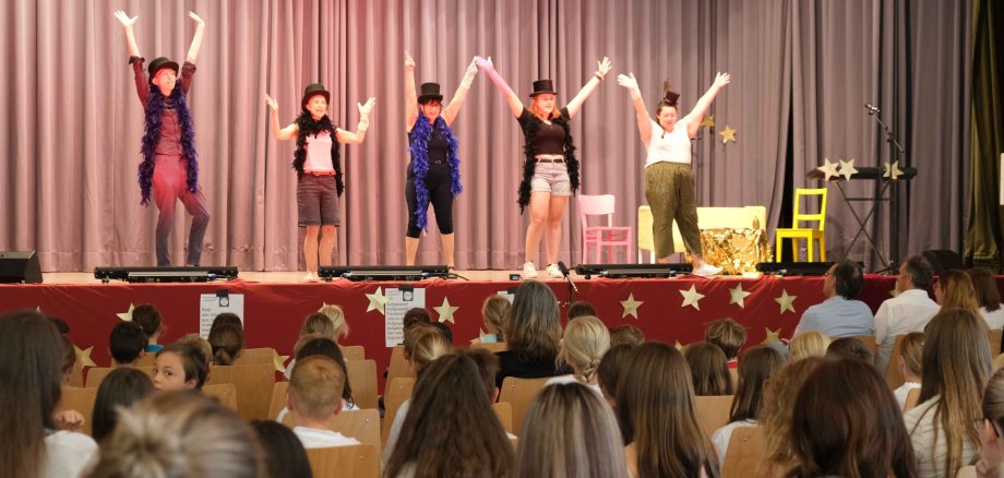 Fünf Darstellerinnen und Darsteller auf einer Bühne. Alle recken die Arme nach oben und tragen einen schwarzen Zylinder. Die Bühne selbst ist an der Stirnseite mit einem roten Tuch bedeckt, auf dem goldene Sterne kleben. Im Vordergrund ist ein Publikum auf Stühlen zu sehen, in dem vorrangig Kinder und Jugendliche sitzen.