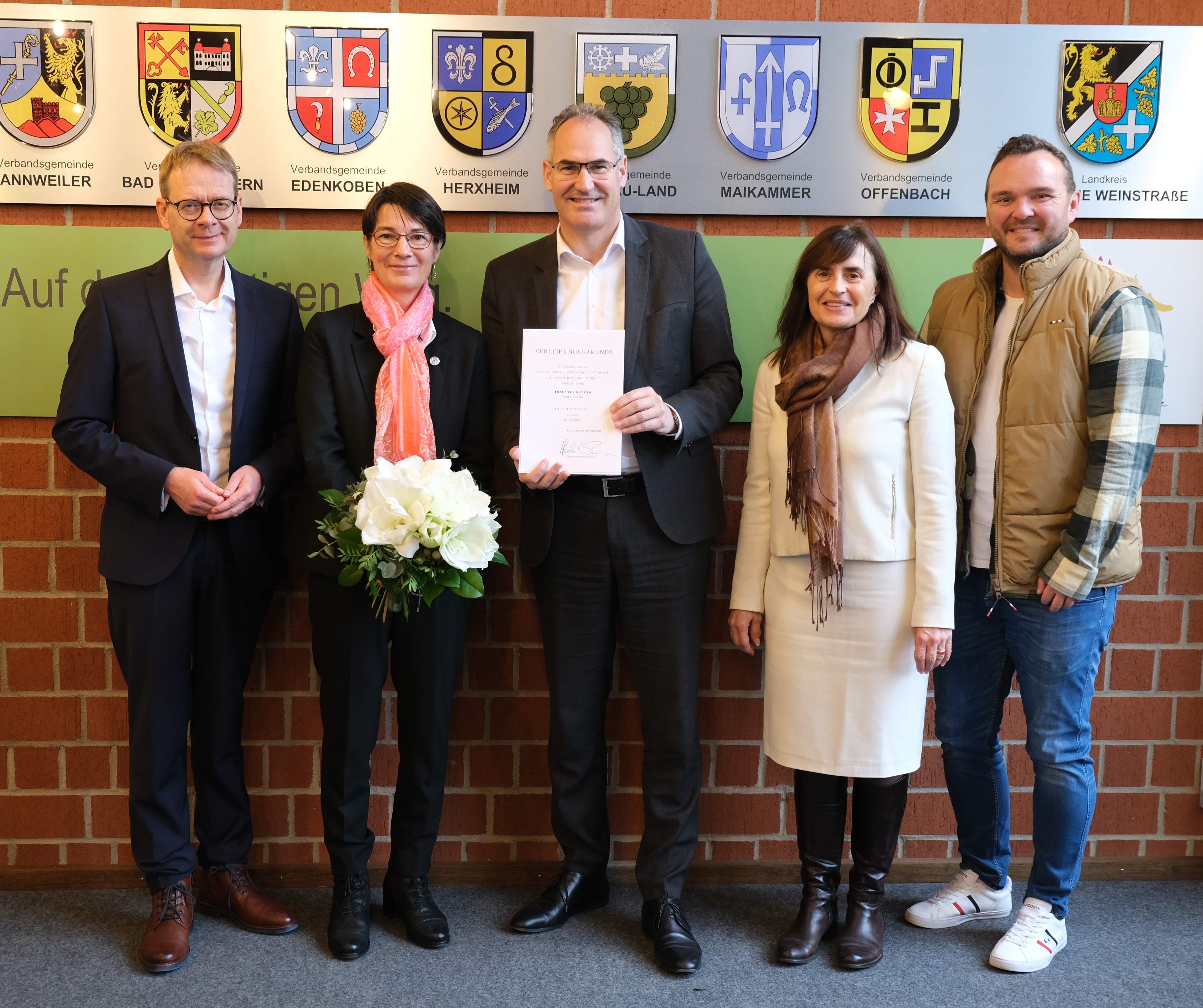 Ute Hormuth (zweite von links) wurde mit der Ehrennadel des Landes Rheinland-Pfalz ausgezeichnet. Ihr gratulierten (von links): Bundestagsabgeordneter Dr. Thomas Gebhart, Landrat Dietmar Seefeldt, Bürgermeisterin Gabriele Flach (Verbandsgemeinde Maikammer) und St. Martins Ortsbürgermeister Timo Glaser. Ute Hormuth hält einen Blumenstrauß, während Dietmar Seefeldt die Urkunde präsentiert. Im Hintergrund sind Wappen der Verbandsgemeinden zu sehen. Foto: KV SÜW. (unterstützt von KI)
