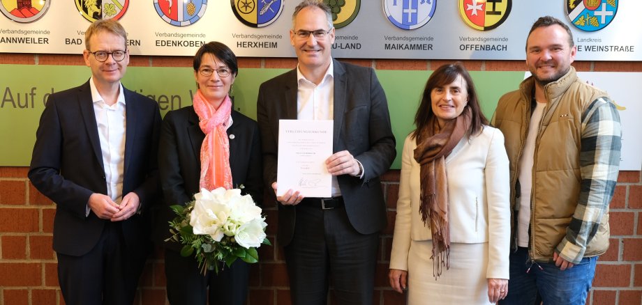 Ute Hormuth (zweite von links) wurde mit der Ehrennadel des Landes Rheinland-Pfalz ausgezeichnet. Ihr gratulierten (von links): Bundestagsabgeordneter Dr. Thomas Gebhart, Landrat Dietmar Seefeldt, Bürgermeisterin Gabriele Flach (Verbandsgemeinde Maikammer) und St. Martins Ortsbürgermeister Timo Glaser. Ute Hormuth hält einen Blumenstrauß, während Dietmar Seefeldt die Urkunde präsentiert. Im Hintergrund sind Wappen der Verbandsgemeinden zu sehen. Foto: KV SÜW. (unterstützt von KI)