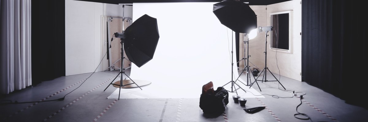 Professionelles Fotostudio mit Beleuchtungs-Setup und weißen Hintergrundwänden. Mehrere Studioleuchten und Softboxen sind um eine leere Fläche herum positioniert, bereit für ein Fotoshooting. Im Vordergrund befinden sich Kabel und eine Tasche mit Ausrüstung, während im Hintergrund große Softboxen und Lichtquellen an der Decke befestigt sind (generiert mit Hilfe von KI).