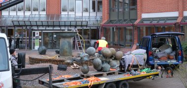 Im Vordergrund liegt die Traube aus Bronze auf einem Kfz-Anhänger, im Hintergrund arbeitet ein Mann am Brunnen, links steht ein Mann unter einem Autokran.
