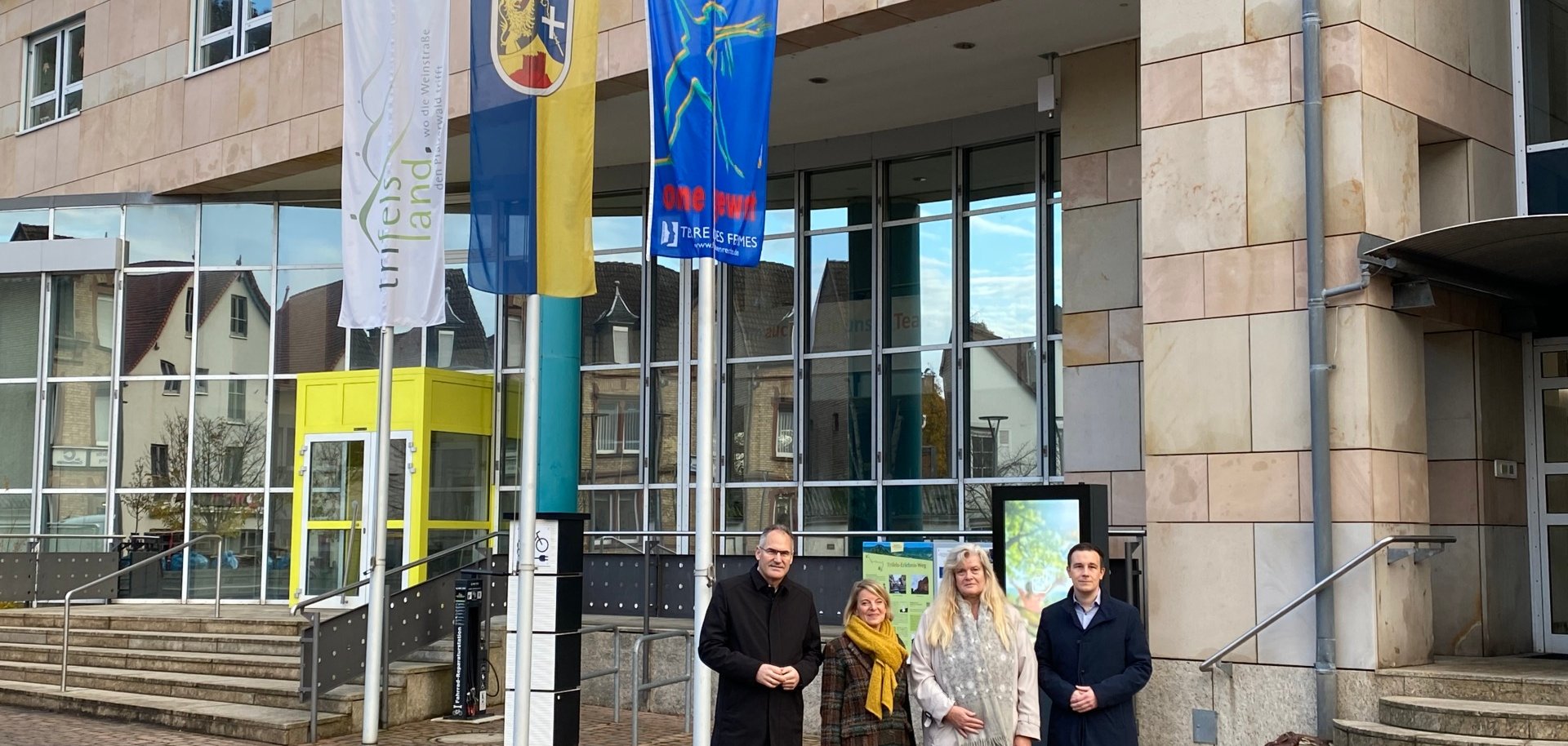 Vier Personen – Landrat Dietmar Seefeldt, SÜW-Gleichstellungsbeauftragte Isabelle Stähle, Gleichstellungsbeauftragte Inge Keller und Bürgermeister Christian Burkhart – stehen vor dem Eingang eines modernen Verwaltungsgebäudes mit Glasfassade. Neben ihnen sind drei Fahnenmasten mit gehissten Flaggen zu sehen, darunter eine blaue Flagge mit der Aufschrift "frei leben – ohne Gewalt" zum Aktionstag gegen Gewalt an Frauen.