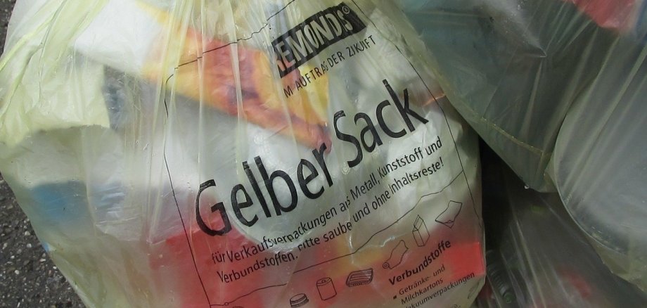 Ein mit Leichtverpackungen gefüllter gelber Sack lehnt an eine Hauswand.