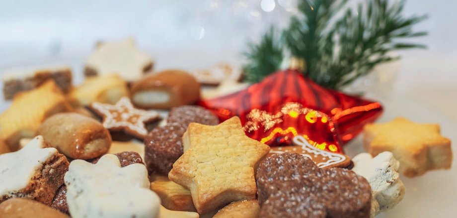 Verschiedene Sorten Weihnachtsplätzchen liegen auf einem Teller, im Hintergrund eine sternförmige Weihnachtskugel sowie ein Tannenzweig.