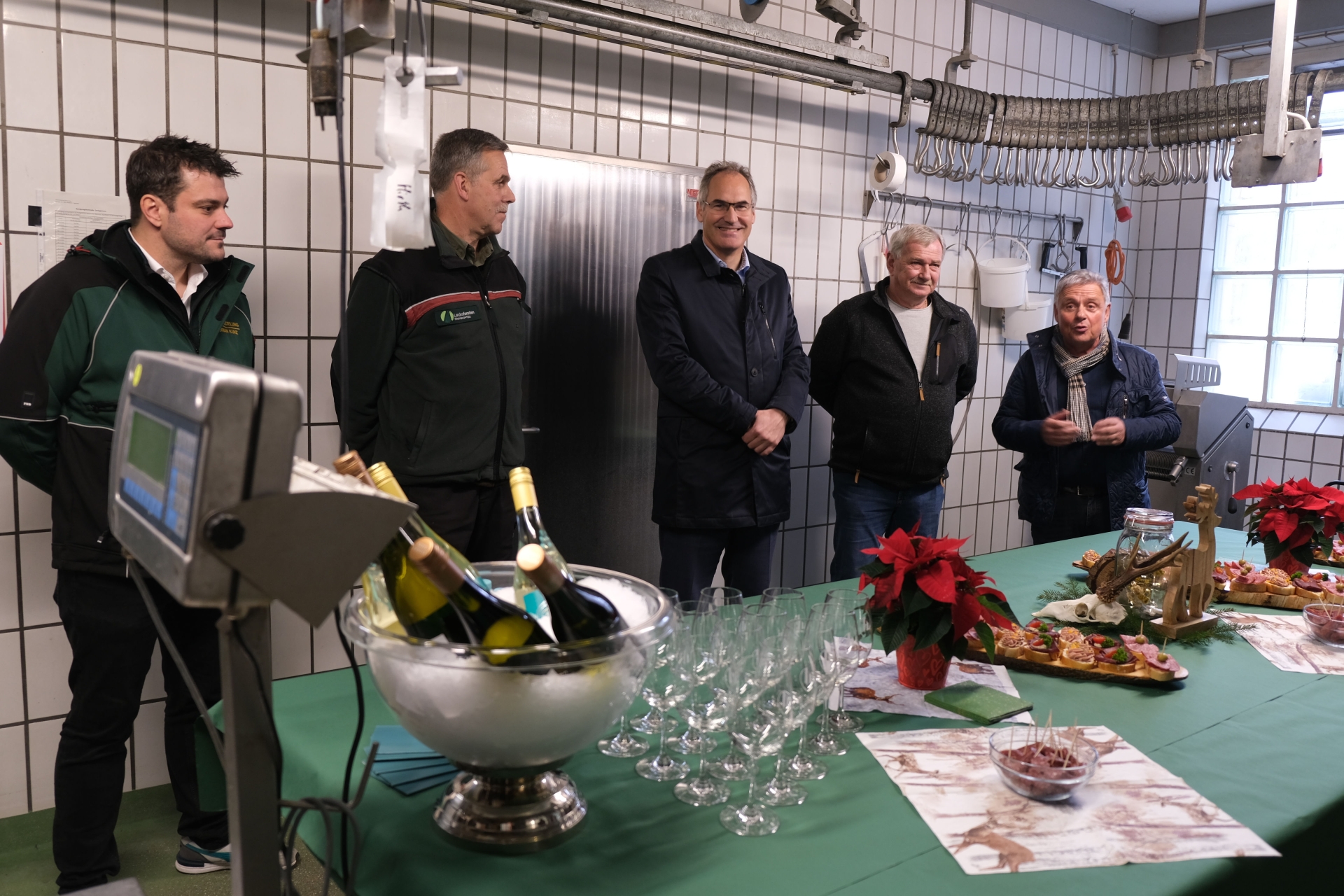 Gruppenbild in der Wildmetzgerei: Inhaber Florian Kunz, Kreisjagdmeister Jörg Sigmund, Landrat Dietmar Seefeldt, Fritz Rück (Obmann für Wildbretmarketing) und Dietmar Pfister, Ortsbürgermeister von Billigheim-Ingenheim. Der Raum ist weiß gefliest, von der Decke hängt eine Schiene mit Fleischhaken. Im Vordergrund vor den abgebildeten Personen steht ein Tisch, auf dem ein Weinkühler mit Eiswürfeln und mehreren Flaschen Wein, Weingläser, Weihnachtssterne und einige Schnittchen stehen. Auf dem tisch liegt eine grüne Tischdecke. Links im Vordergrund ist außerdem eine Waage zu sehen.