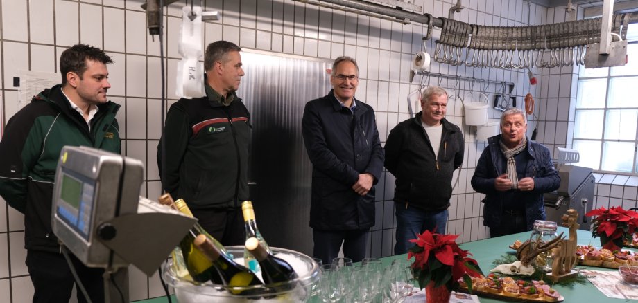 Gruppenbild in der Wildmetzgerei: Inhaber Florian Kunz, Kreisjagdmeister Jörg Sigmund, Landrat Dietmar Seefeldt, Fritz Rück (Obmann für Wildbretmarketing) und Dietmar Pfister, Ortsbürgermeister von Billigheim-Ingenheim. Der Raum ist weiß gefliest, von der Decke hängt eine Schiene mit Fleischhaken. Im Vordergrund vor den abgebildeten Personen steht ein Tisch, auf dem ein Weinkühler mit Eiswürfeln und mehreren Flaschen Wein, Weingläser, Weihnachtssterne und einige Schnittchen stehen. Auf dem tisch liegt eine grüne Tischdecke. Links im Vordergrund ist außerdem eine Waage zu sehen.