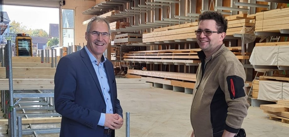Landrat Dietmar Seefeldt (links) und Alexander Wissing, Geschäftsführer eines Zimmereibetriebs und Sägewerks, stehen in einer großen, lichtdurchfluteten Firmenhalle mit Regalen voller Holzmaterialien in Kapellen-Drusweiler. (unterstützt von KI)
