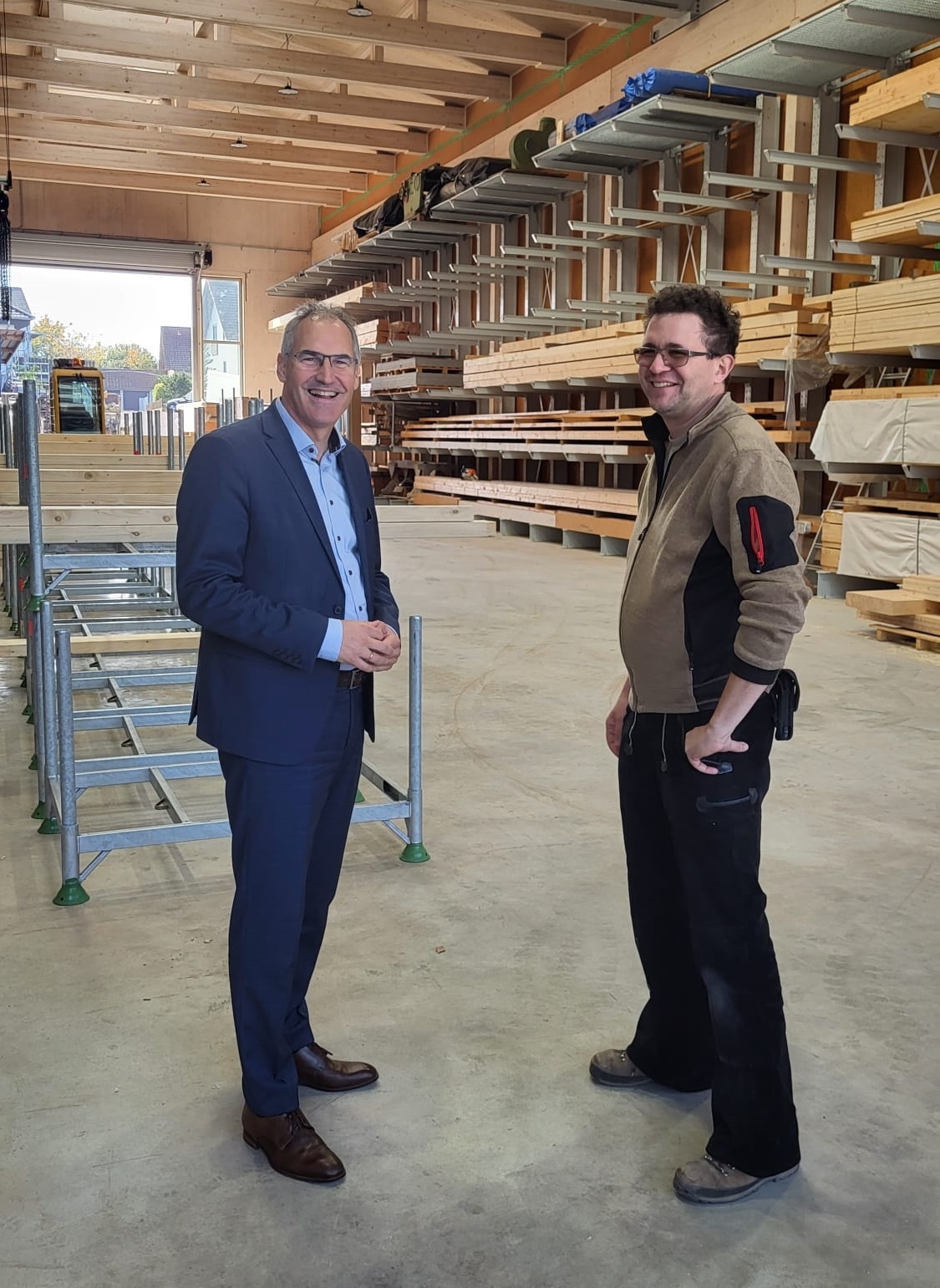 Landrat Dietmar Seefeldt (links) und Alexander Wissing, Geschäftsführer eines Zimmereibetriebs und Sägewerks, stehen in einer großen, lichtdurchfluteten Firmenhalle mit Regalen voller Holzmaterialien in Kapellen-Drusweiler. (unterstützt von KI)