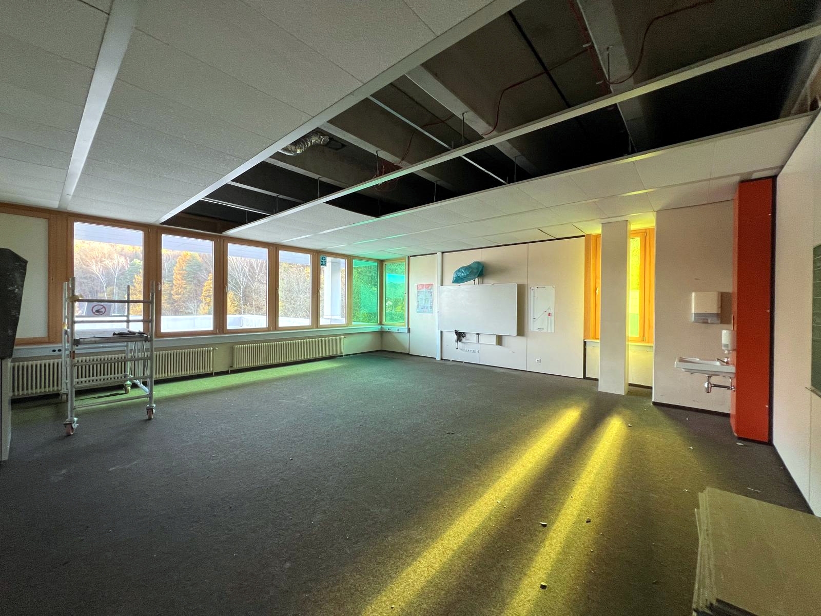 Ein leerer Unterrichtsraum im Staufer-Schulzentrum in Annweiler mit freigelegten Decken und abgebauten Einrichtungsgegenständen. Durch die großen Fenster fällt Sonnenlicht, das den Raum erhellt. Im Hintergrund sind herbstlich gefärbte Bäume zu sehen, während im Raum Bauarbeiten sichtbar vorbereitet sind.