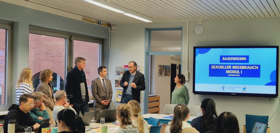 Menschen in einem Klassenraum vor einer digitalen Tafel.