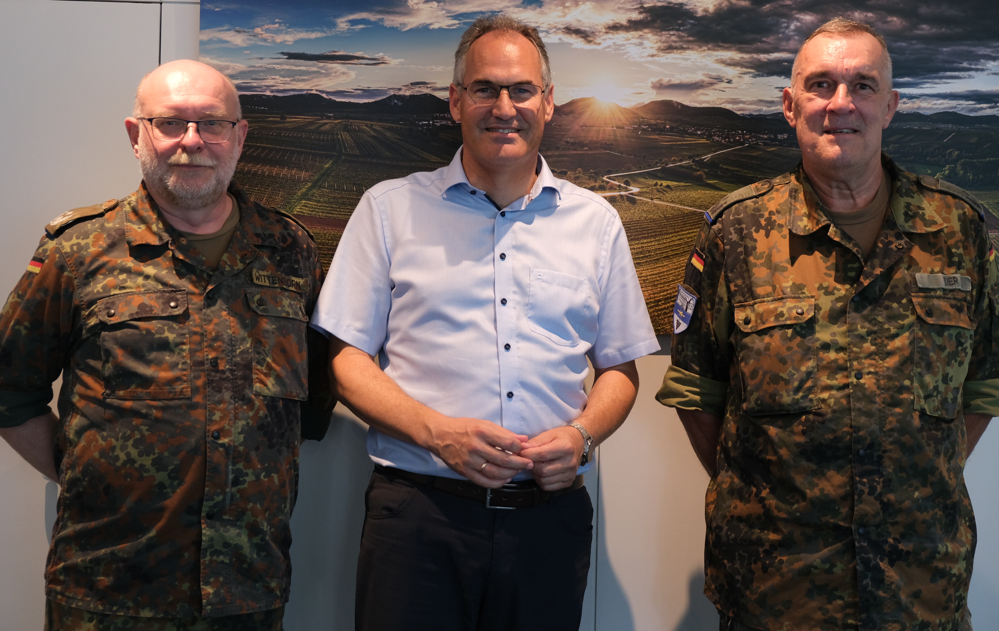 Landrat Dietmar Seefeldt empfängt die Oberstleutnante der Bundeswehr Kay Wittenborn und Wolfgang Eder im Kreishaus. Auf dem Bild stehen (von links nach rechts) die zwei uniformierten Oberstleutnante der Bundeswehr neben Landrat Seefeldt, der ein helles Hemd trägt. Im Hintergrund ist eine malerische Landschaft mit Feldern und einem Sonnenuntergang zu sehen, die auf einem Wandbild abgebildet ist. Die beiden Bundeswehrangehörigen tragen typische Bundeswehr-Tarnuniformen und die deutschen Flaggenabzeichen auf ihren Ärmeln.