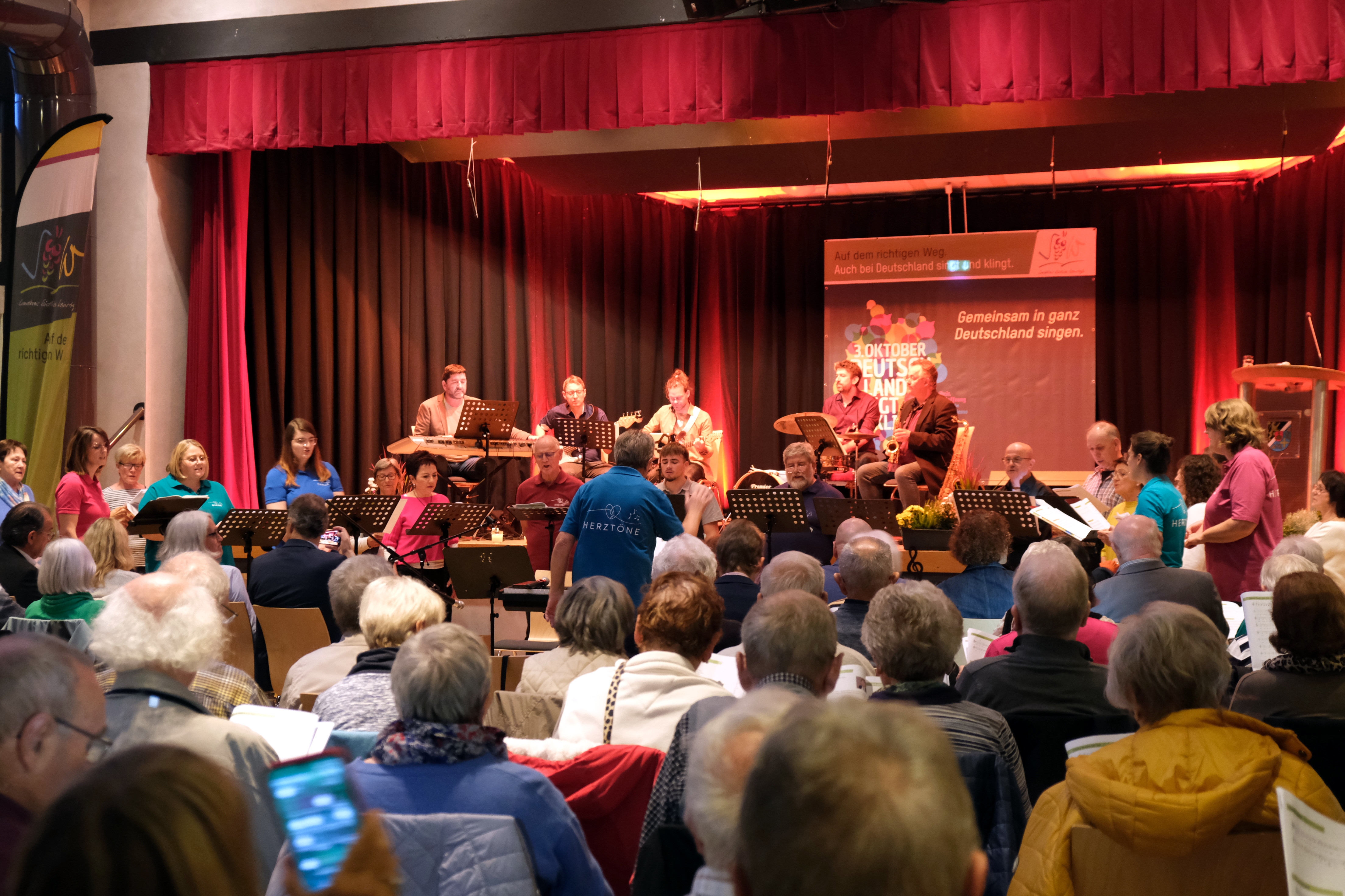 Auf der Bühne des Dorfgemeinschaftshauses Bornheim spielt die Band "SÜW Cuvée" auf ihren Instrumenten, davor singen Chormitglieder des Chors "Herztöne". Im Vordergrund viele Menschen auf Sitzplätzen.