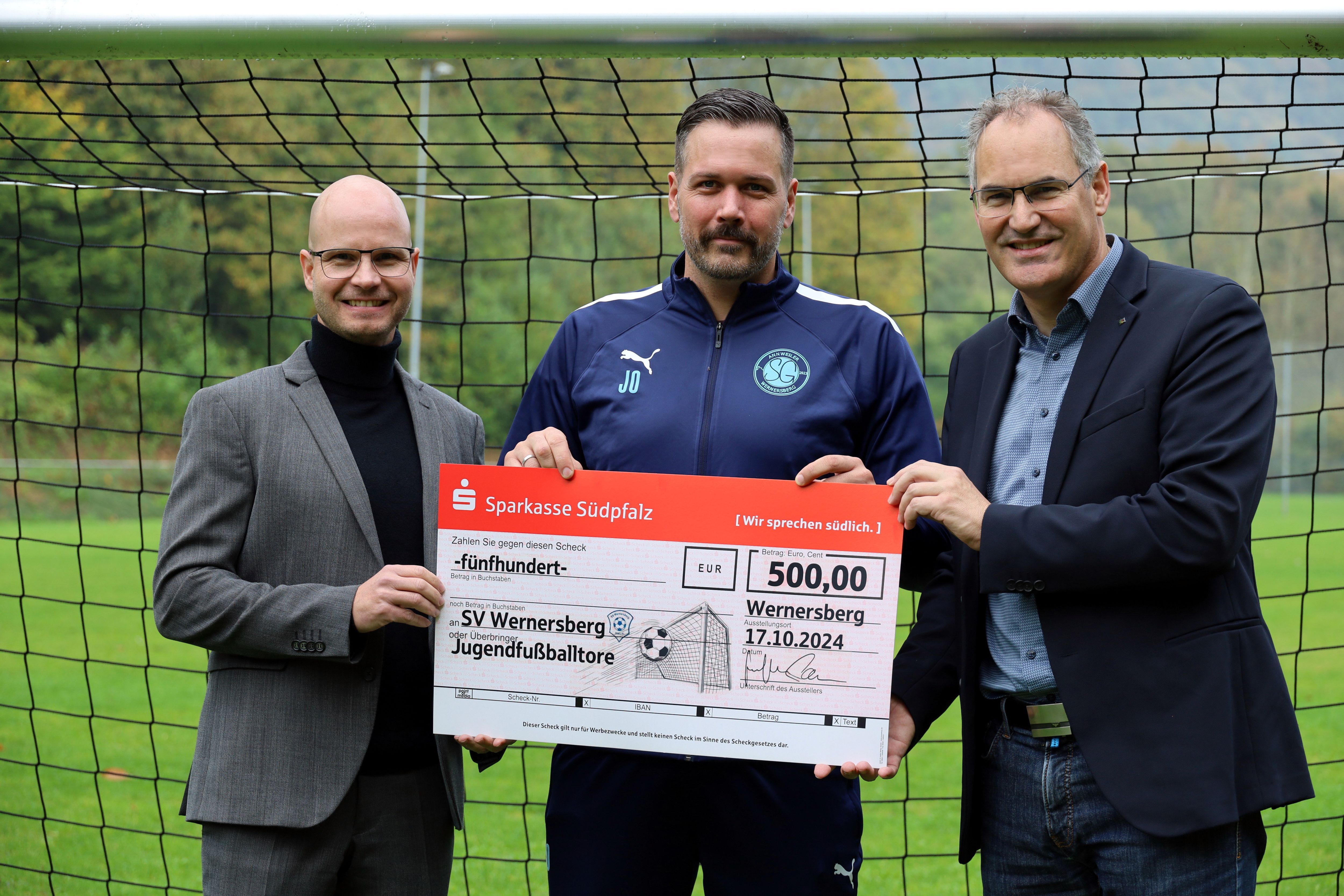 Sparkassenvorsitzender Svend Larsen, Jan Ohlert vom SV Wernersberg und Landrat Dietmar Seefeldt stehen vor einem Fußballtor und halten einen großen symbolischen Scheck der Sparkasse Südpfalz über 500 Euro in die Kamera. Der Scheck ist an den SV Wernersberg für die Unterstützung der Jugendarbeit im Fußballverein adressiert. Hintergrund zeigt ein grünes Fußballfeld und eine hügelige Landschaft.