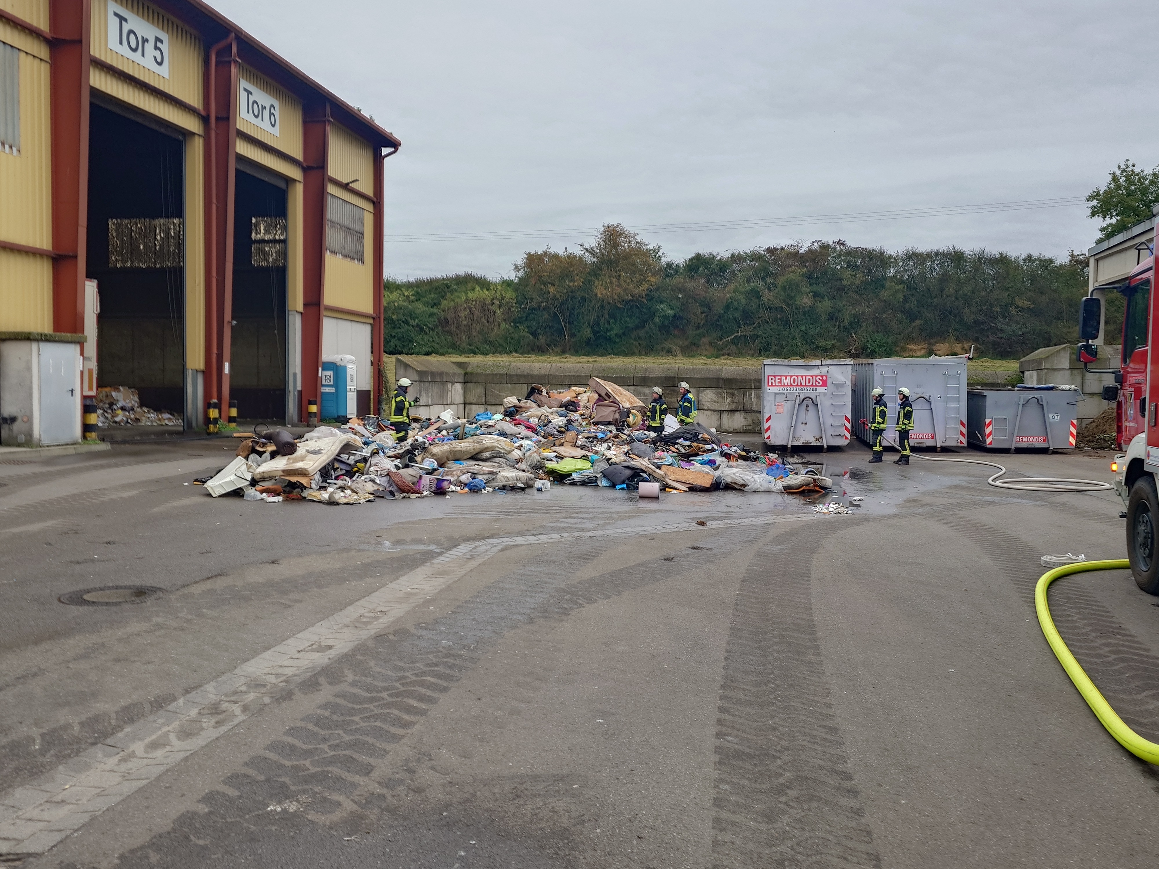 Feuerwehrleute stehen um einen großen Müllhaufen auf einer betonierten Fläche.