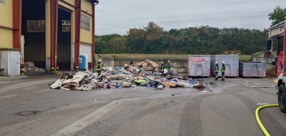 Feuerwehrleute stehen um einen großen Müllhaufen auf einer betonierten Fläche.