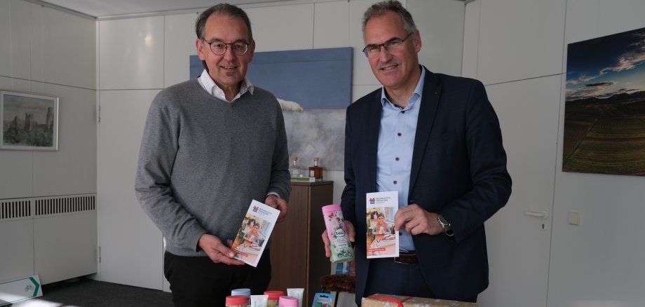 Landrat Dietmar Seefeldt und Erster Kreisbeigeordneter Georg Kern stehen in einem Büro und halten Flyer zur Aktion Weihnachtspäckchenkonvoi in den Händen. Auf dem Tisch vor ihnen sind bunt verpackte Geschenke, Spielzeug und Pflegeprodukte zu sehen, die für bedürftige Kinder gesammelt wurden.