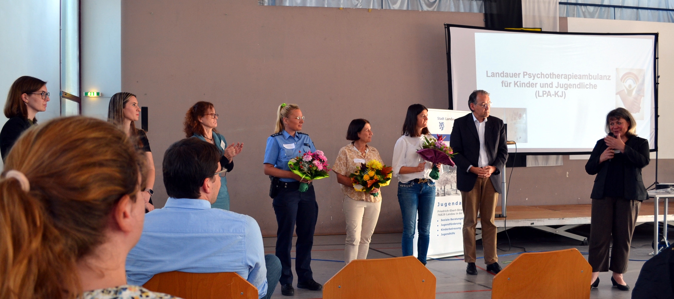 Auf dem Bild stehen mehrere Personen in einer Halle auf einer Bühne. Drei Frauen, darunter eine Polizistin, halten Blumensträuße in den Händen und werden von den umstehenden Personen, darunter auch ein Mann im Anzug, applaudiert. Im Hintergrund ist eine Leinwand zu sehen, auf der die „Landauer Psychotherapieambulanz für Kinder und Jugendliche (LPA-KJ)“ vorgestellt wird. Vor der Bühne sitzen weitere Personen und folgen der Veranstaltung.