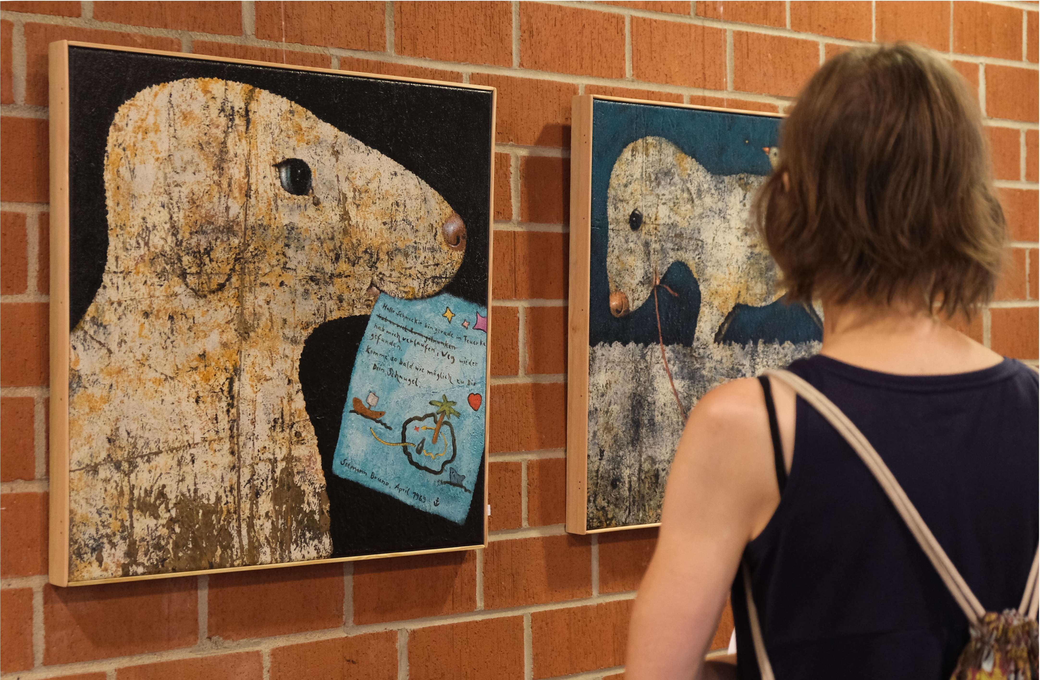 Eine Besucherin betrachtet zwei quadratische Gemälde von Matthias Göhr, die an einer roten Backsteinwand hängen. Das linke Bild zeigt ein Tier, das einem Schaf ähnelt und ein blaues Papier im Maul hält, auf dem ein handschriftlicher Text sowie kleine Illustrationen zu sehen sind. Das rechte Bild zeigt ein ähnliches Tier, das an einer Leine befestigt ist und auf einem dunklen Hintergrund dargestellt wird.