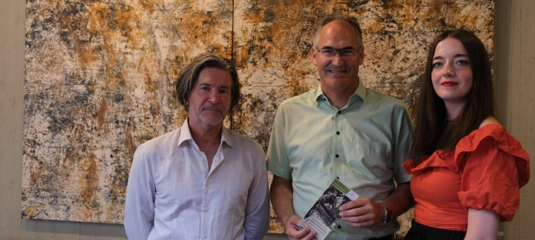 Bei der Vernissage (von links): Künstler Matthias Göhr, Landrat Dietmar Seefeldt und Sabine Adler, die in die Ausstellung einführte. Foto: KV SÜW Der Künstler Matthias Göhr steht zusammen mit Landrat Dietmar Seefeldt und der Kunsthistorikerin Sabine Adler vor einem großformatigen Gemälde. Das Bild im Hintergrund ist abstrakt, mit dominierenden gelblich-braunen und schwarzen Farben. Alle drei Personen lächeln in die Kamera.