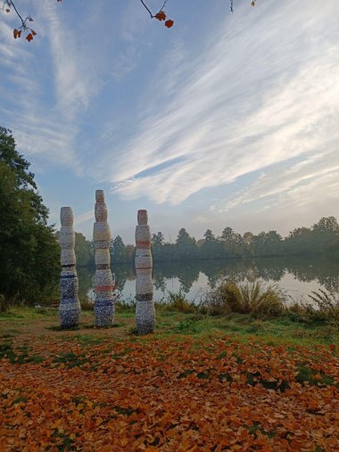 „Three Pillars“, ein Kunstobjekt von Karen van der Molen für den Kirrweiler Kunstpfad. Foto: Tourismusbüro Kirrweiler Drei Pfeiler stehen an einem Seeufer.