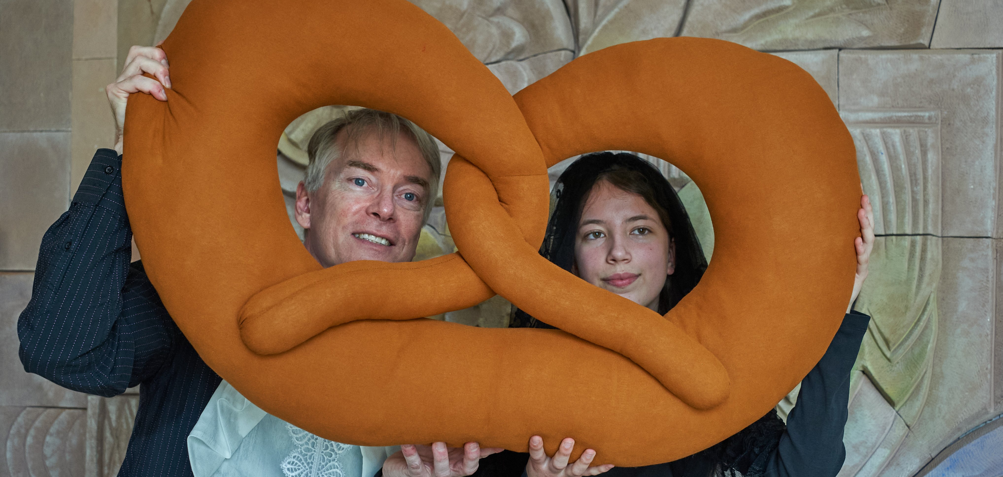 Zwei Personen halten eine übergroße Brezel aus Stoff vor sich, durch deren Löcher sie hindurchschauen. Die linke Person trägt ein Hemd mit einem Spitzenkragen, die rechte Person ist in dunkler Kleidung und mit einem Schleier gekleidet. Beide lächeln leicht in die Kamera.