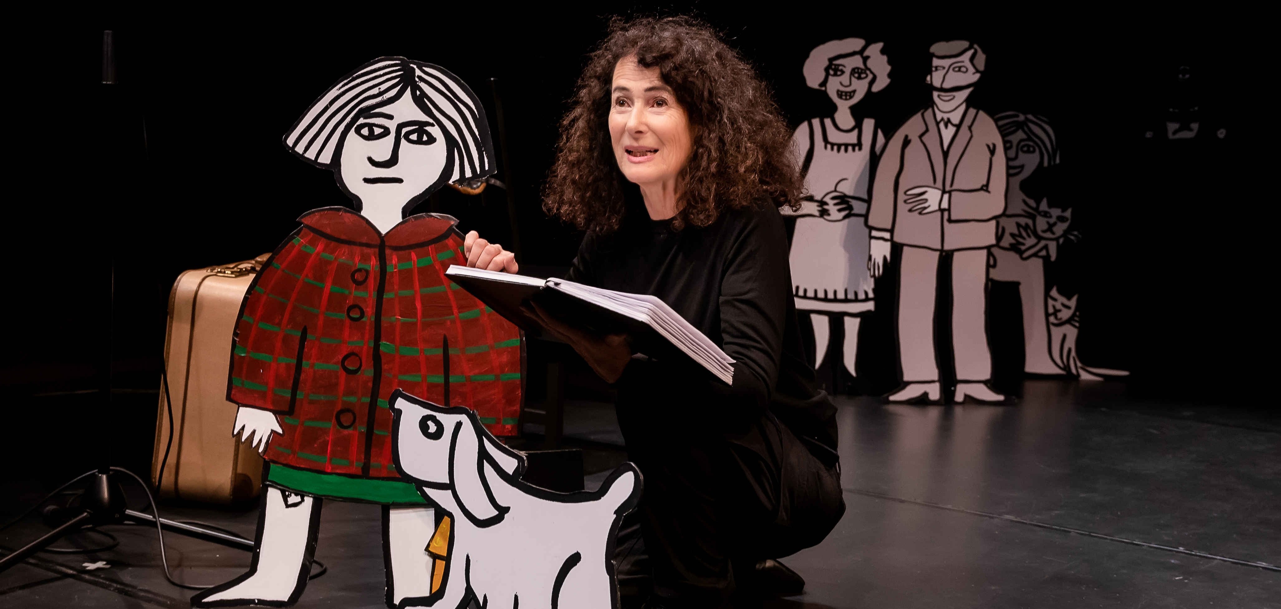 Eine Frau mit lockigem Haar sitzt auf der Bühne vor einer bemalten Pappfigur eines Mädchens in einem roten Mantel mit einem weißen Hund. Die Szene stammt aus dem Theaterstück „All that matters – worauf es ankommt“.