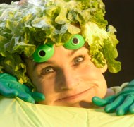 Auch das Kindermusical „FRISCH FROSCH FREI“ ist zu sehen. Foto: Theater Beatrice Hutter. Ein lachender Mensch mit einer Perücke aus Salatblättern und grünen Handschuhen ist zu sehen. Die Person trägt grüne Froschaugen auf dem Kopf. Diese humorvolle Szene stammt aus dem Kindermusical „FRISCH FROSCH FREI“.