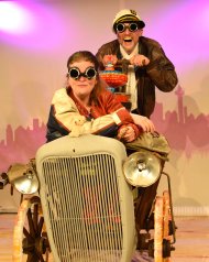 Chaotisch und humorvoll geht es zu in „Copper, Flint und der Fabulotor“. Foto: Heinz Kindler Zwei Schauspieler in einem fantasievollen Fahrzeug. Einer der Darsteller, verkleidet als Kapitän mit weißer Mütze, lacht im Hintergrund, während die vordere Figur mit Fliegerbrille nach vorne blickt. Die Szene stammt aus dem chaotisch-humorvollen Stück „Copper, Flint und der Fabulotor“.