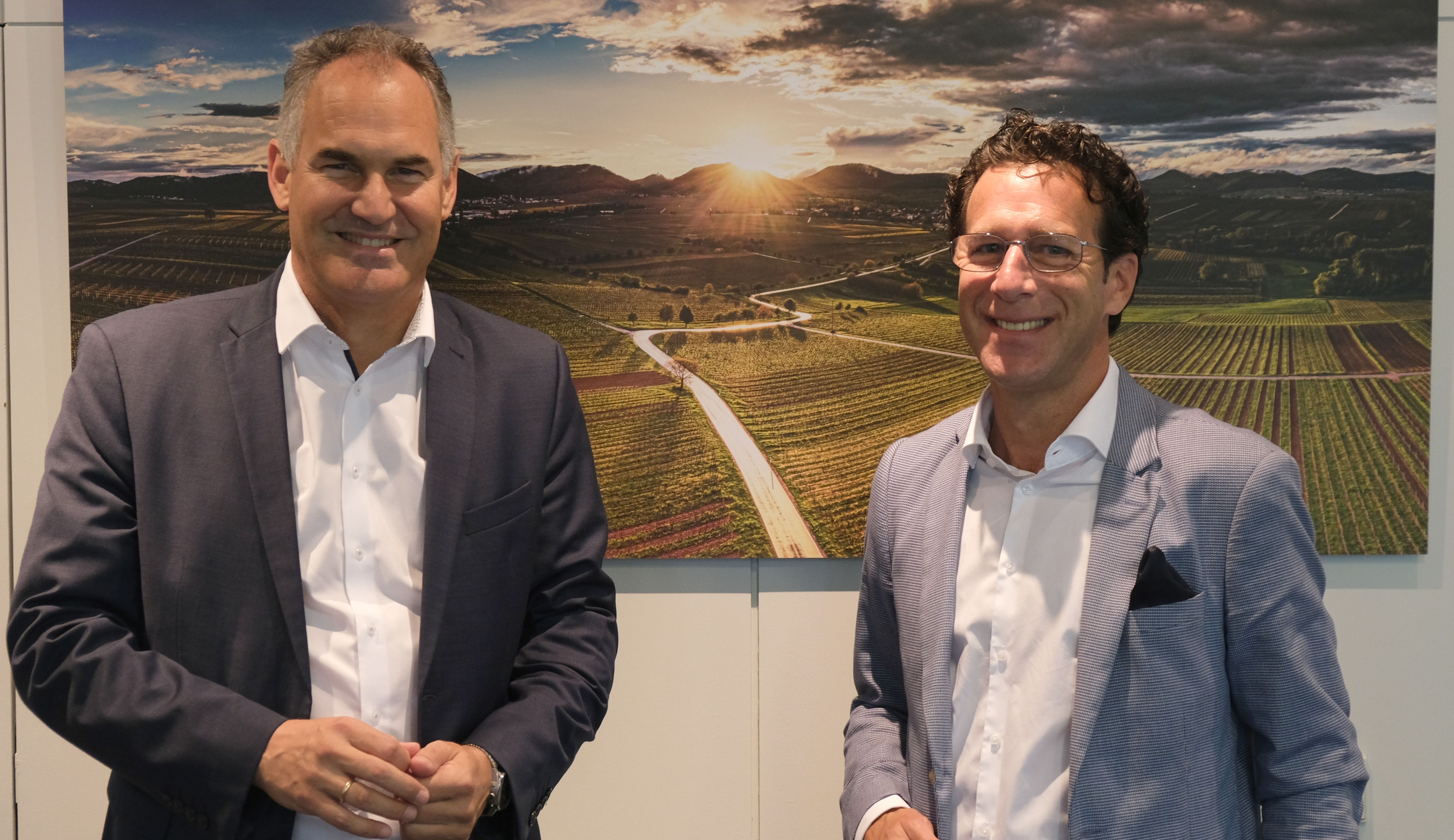 Zwei Männer in Anzügen stehen vor einem Foto der Weinbergslandschaft der Südlichen Weinstraße.