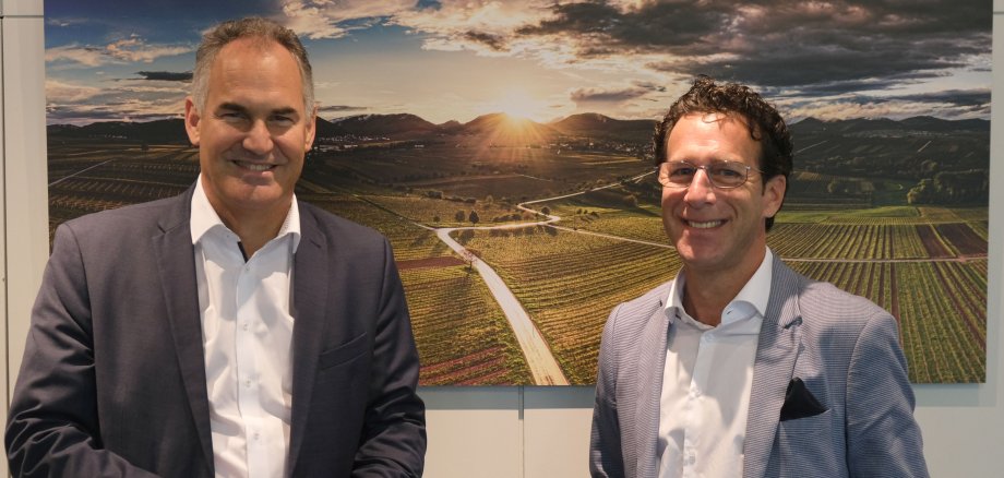 Zwei Männer in Anzügen stehen vor einem Foto der Weinbergslandschaft der Südlichen Weinstraße.