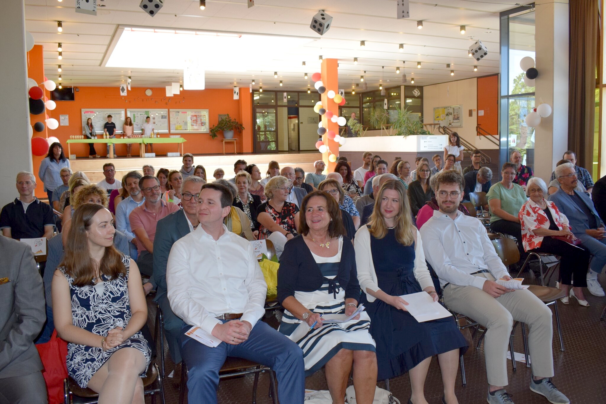 „Gruppenfoto der Verabschiedung von Ingrid Baumgartner-Schmitt, umgeben von zahlreichen Gästen, die in mehreren Reihen sitzen und stehen. Baumgartner-Schmitt sitzt in der Mitte der vorderen Reihe und lächelt in die Kamera.“