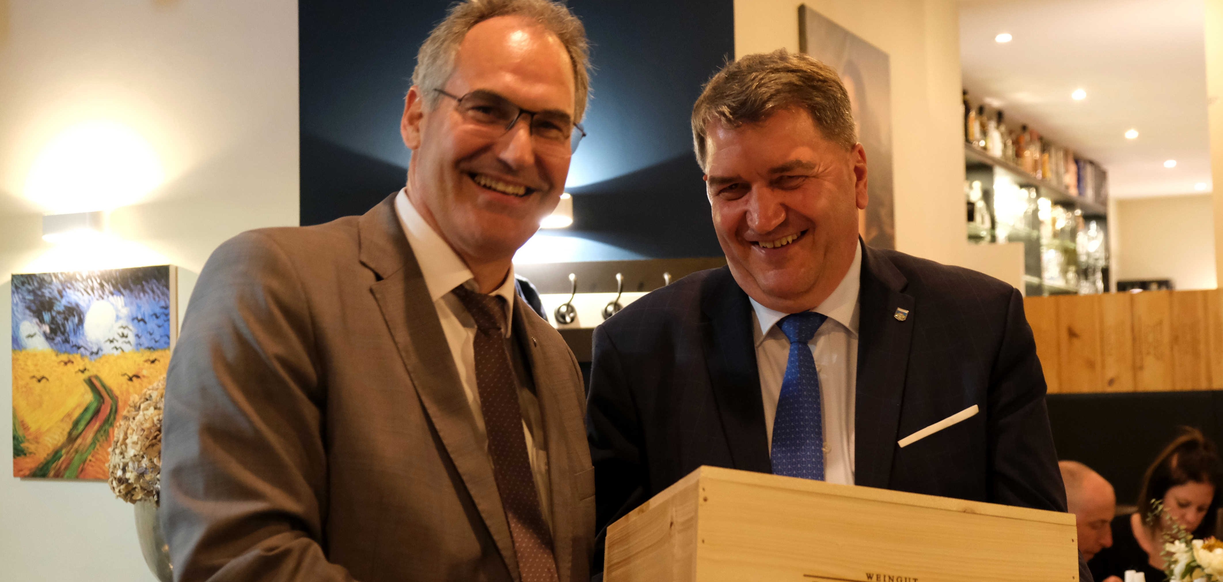 Landrat Dietmar Seefeldt (links) überreicht dem Stadtpräsidenten von Oświęcim, Janusz Chwierut (rechts), eine Holzkiste vom Weingut Dr. Wehrheim aus Birkweiler. Beide Männer lächeln und stehen in einem stilvoll beleuchteten Raum mit modernen Kunstwerken an den Wänden. Die Übergabe symbolisiert die freundschaftliche Verbundenheit zwischen dem Landkreis Südliche Weinstraße und der Stadt Oświęcim.